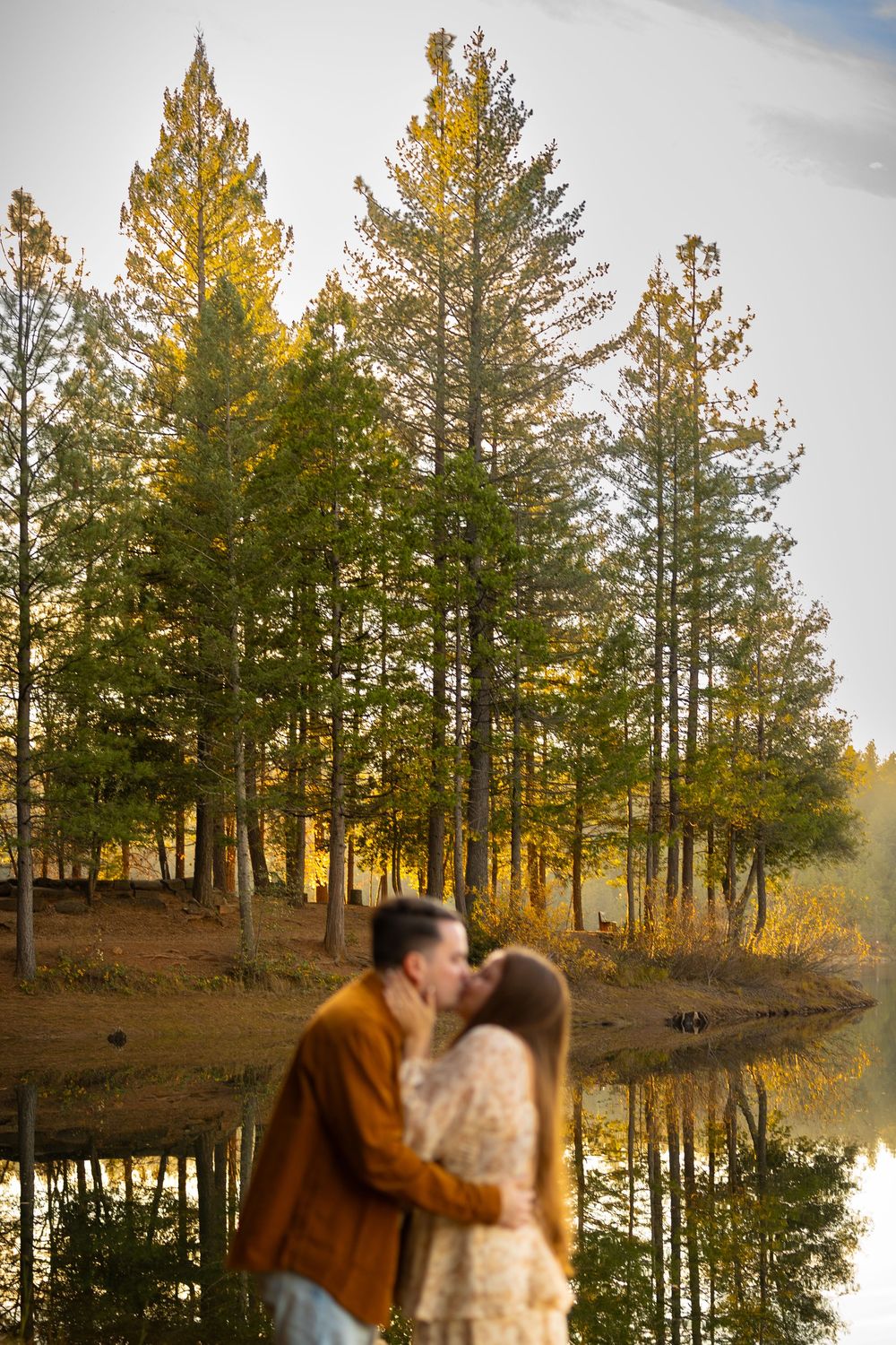 A couple poses romantically against a scenic lake with tall pine trees reflecting in the water at sunset.