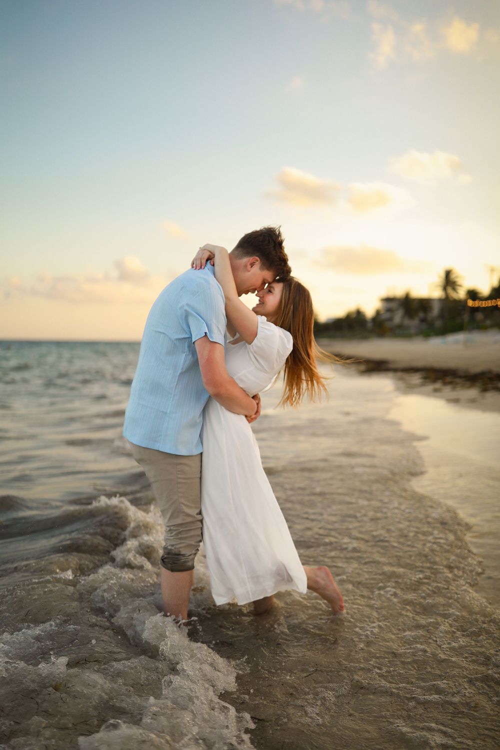 A couple shares a romantic moment while standing in ocean waves at sunset.