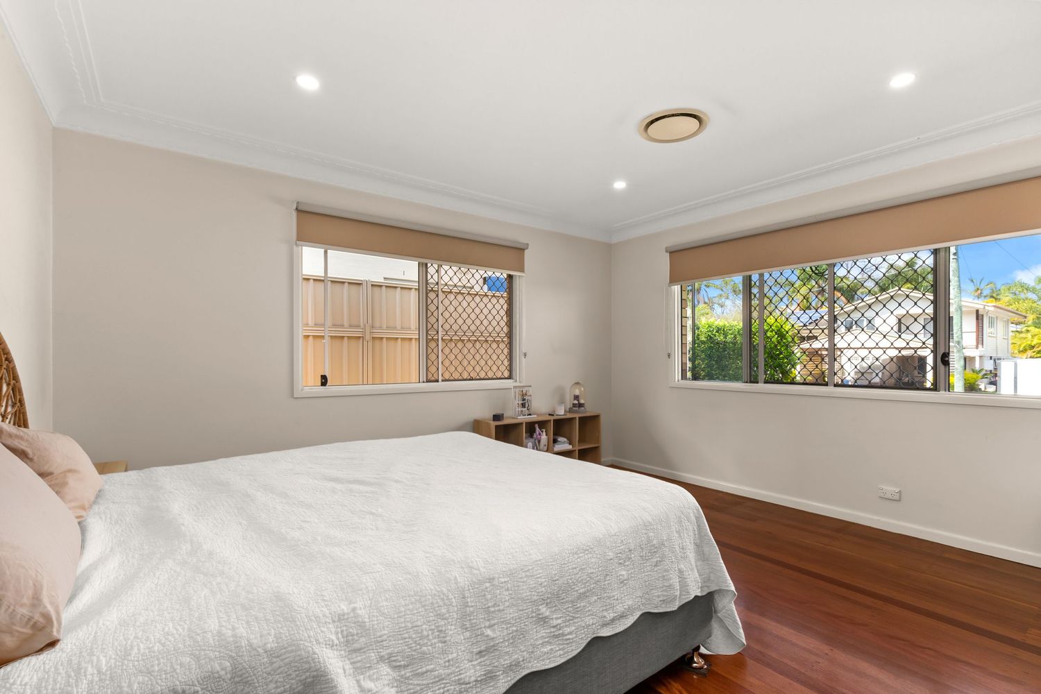 Sunlit bedroom with dark hardwood floors, white bedding, roller blinds, and lattice fence views through windows.