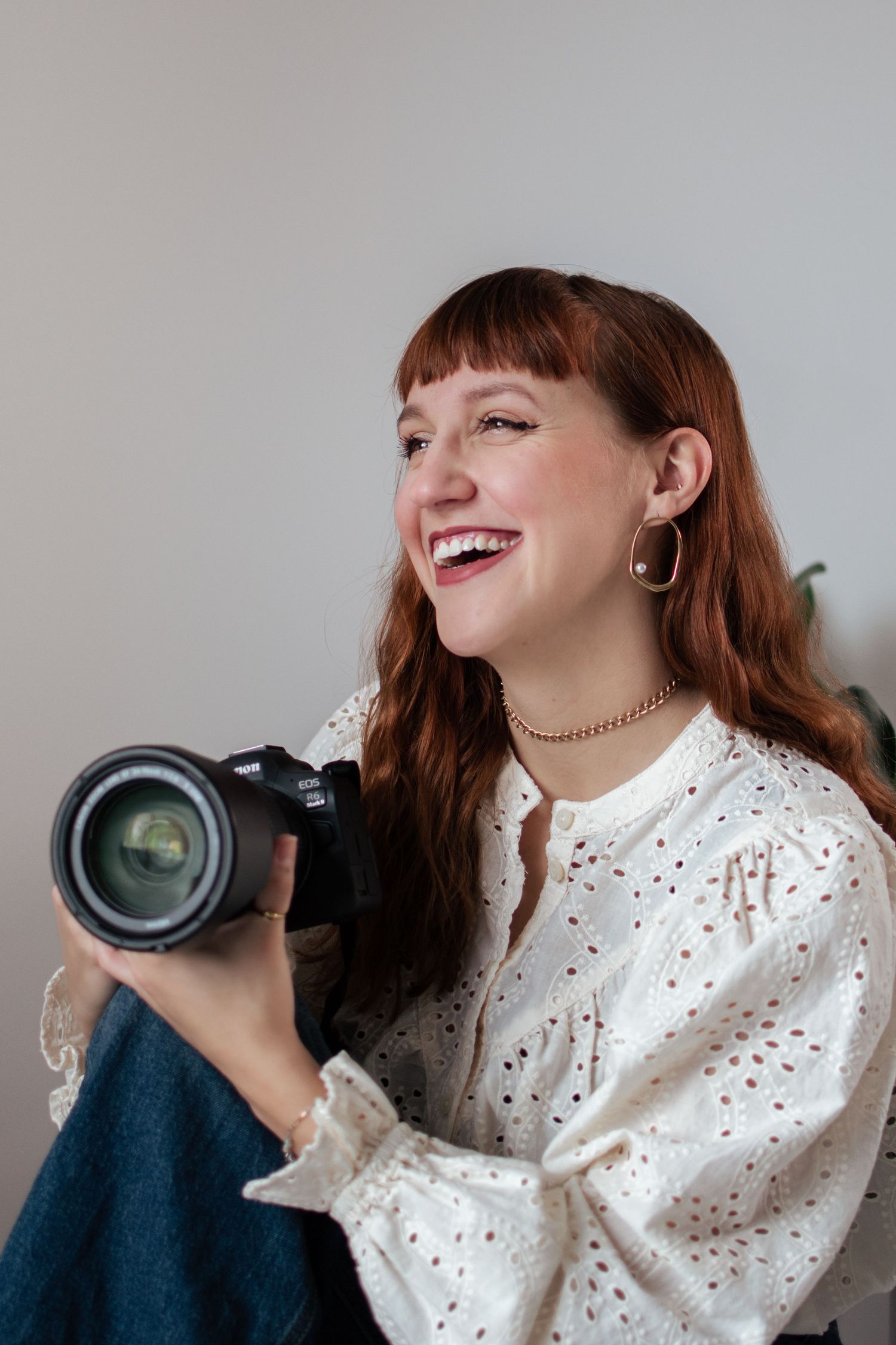 Professional photographer in white polka dot blouse laughs while holding camera lens during creative photoshoot session.