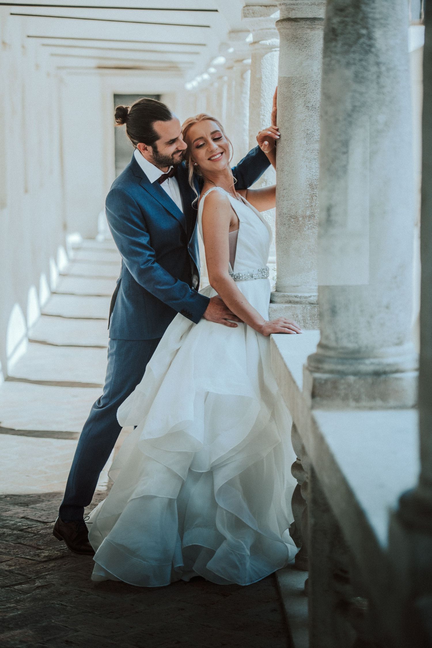 Romantic wedding portrait of couple in formal attire posing in a columned corridor.