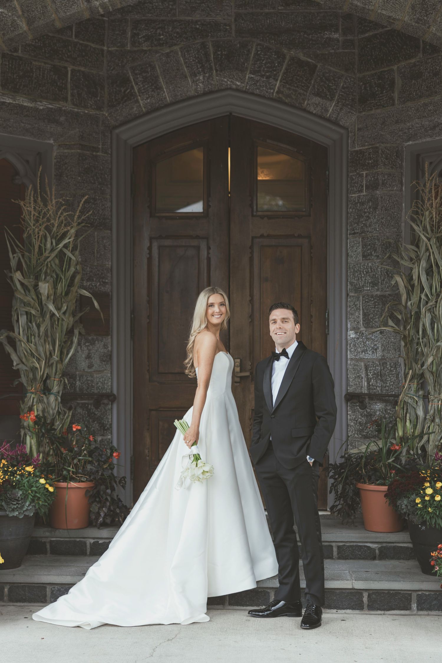 A bride and groom stand together on a stone porch with potted plants and flowers near a grand entrance.
