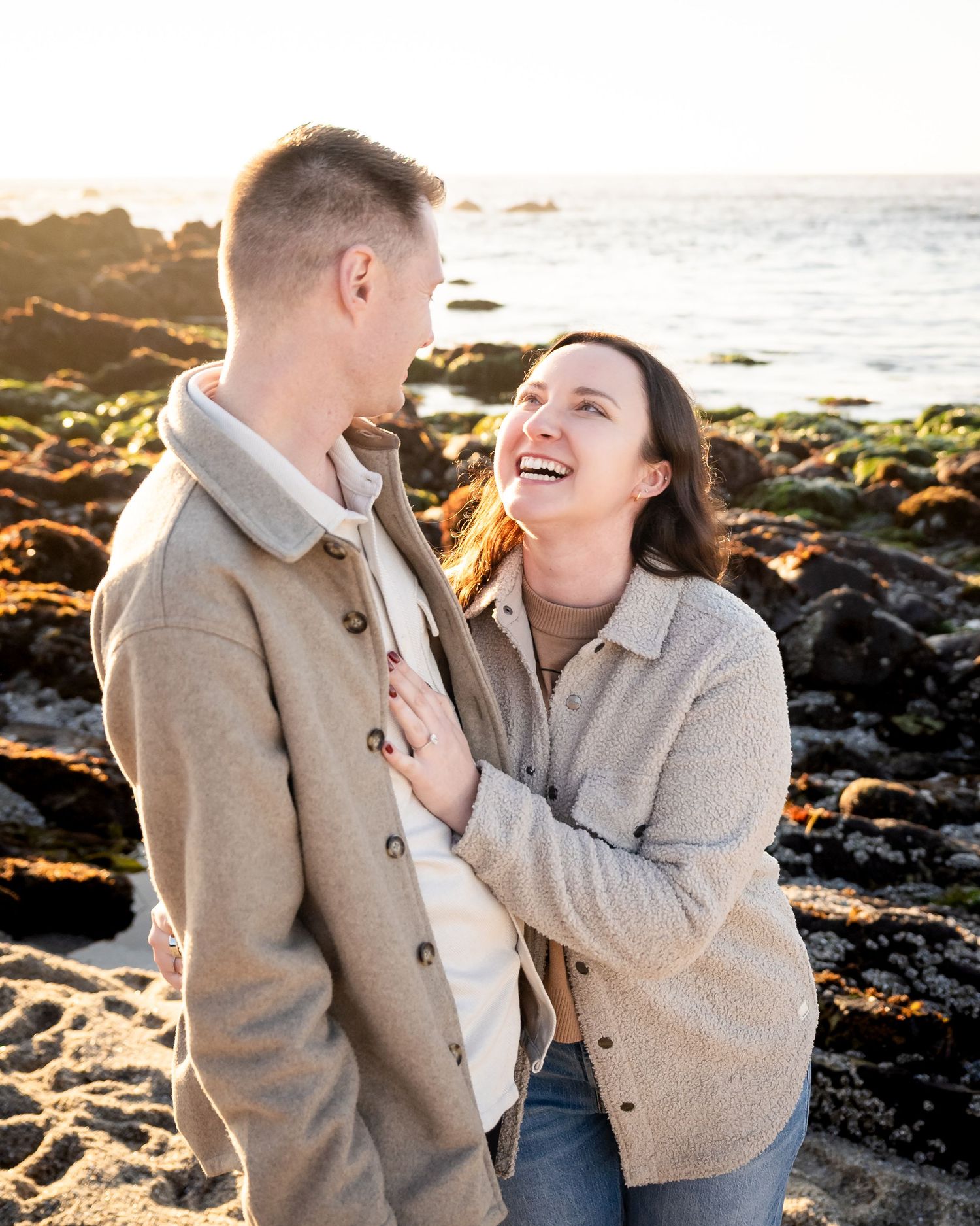A couple shares joyful moments together on a rocky beach at sunset with golden light illuminating their laughter.
