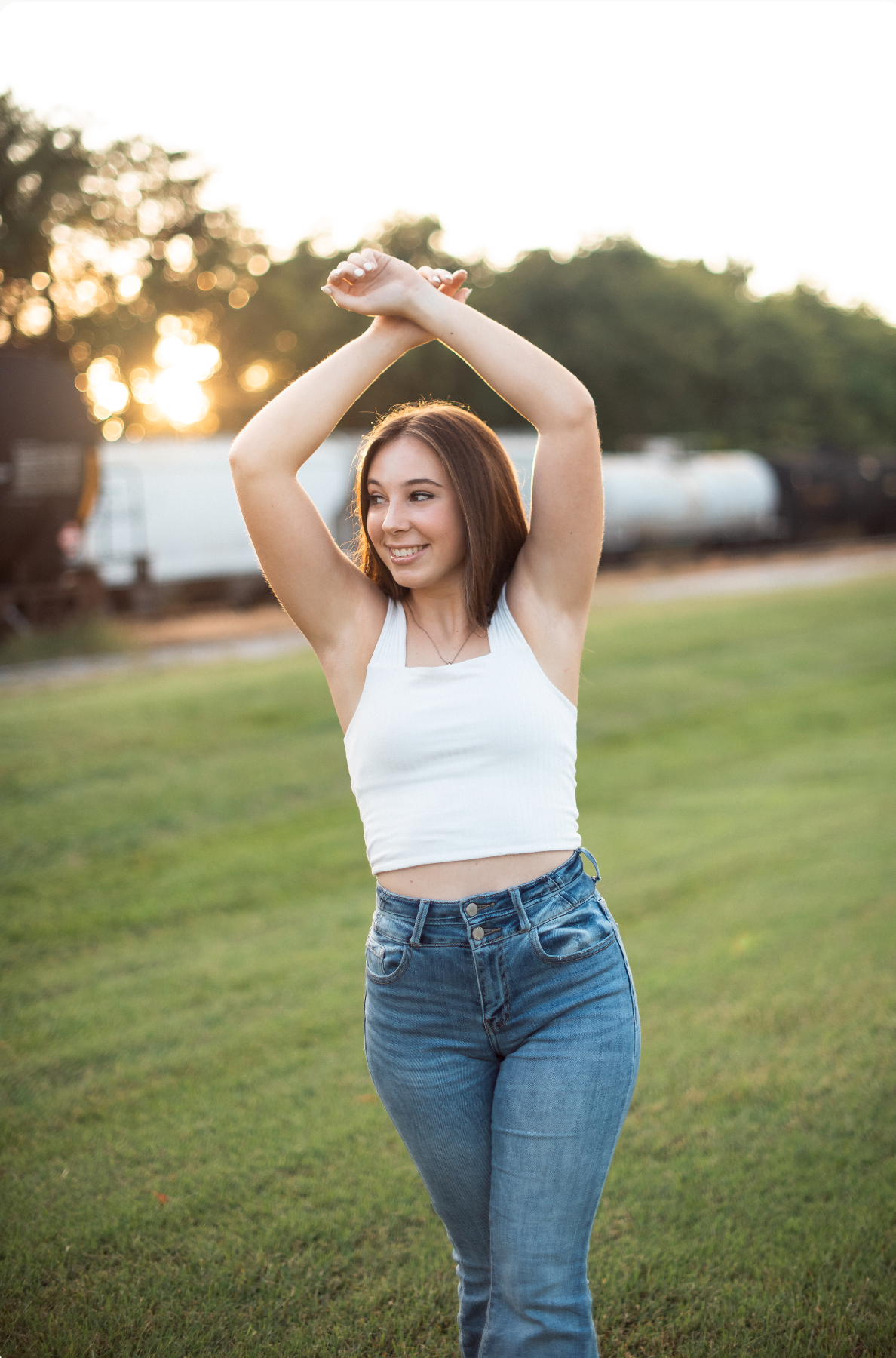 A person in a white crop top and blue jeans poses outdoors at sunset in a grassy field.