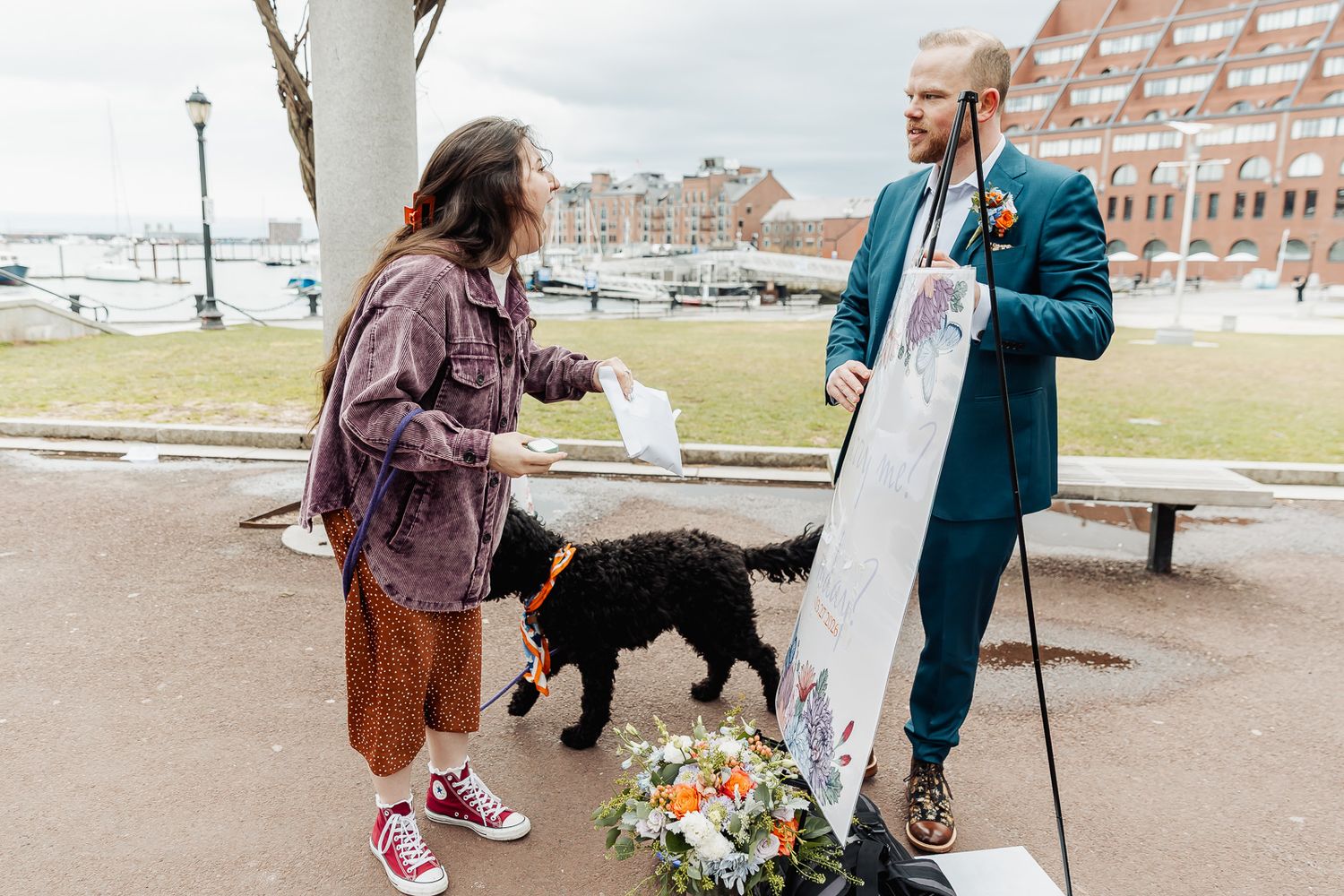 Couple with black dog holding flowers and a sign outdoors near a brick building on a cloudy day.