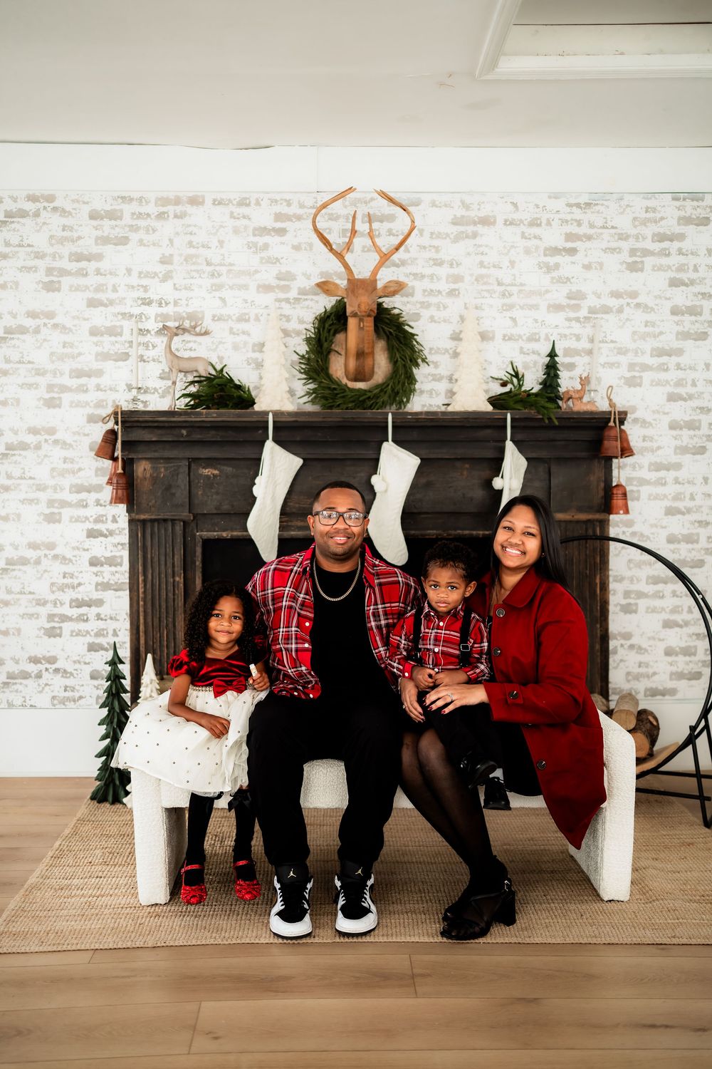 Family of four poses in matching red plaid outfits before a decorated Christmas fireplace with stockings and deer decor.