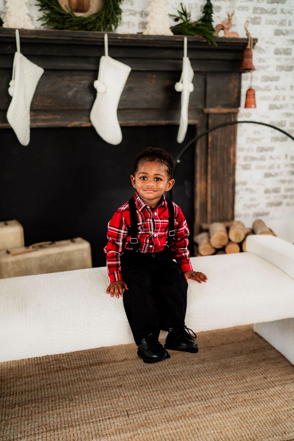 Adorable young boy in red plaid shirt and suspenders sits smiling before a Christmas fireplace with white stockings.