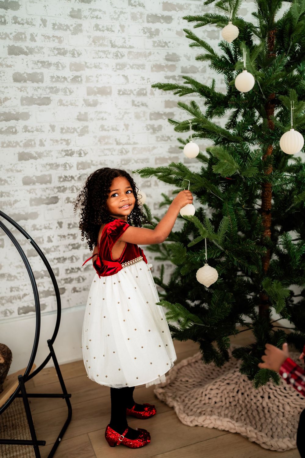Young girl in red and white dress decorating a Christmas tree with white ornaments in a festive room.