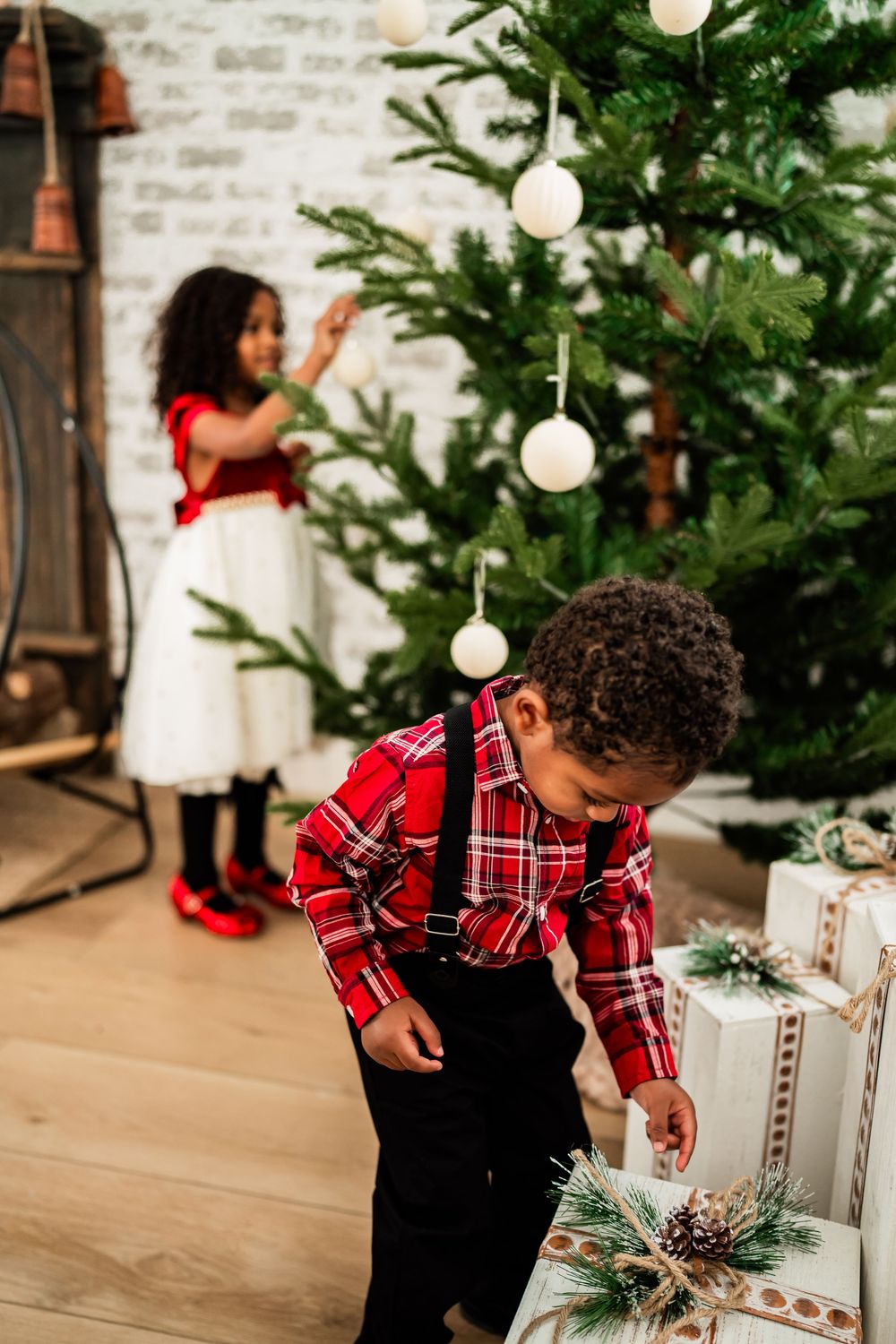 Two African American children decorating a Christmas tree, boy in red plaid shirt with gifts, girl in white dress with red shoes.