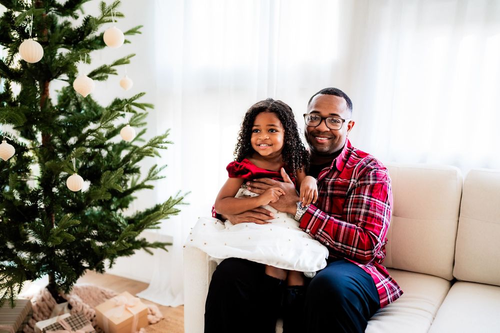 A father and daughter smile together beside a decorated Christmas tree in a bright living room.