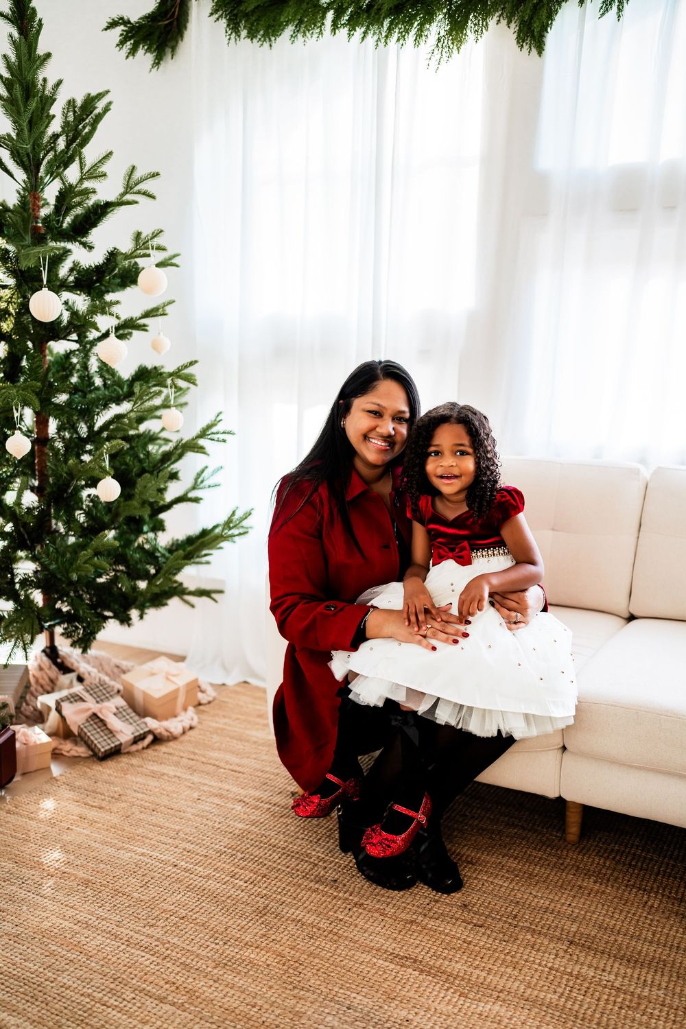 Mother in red dress hugging young daughter in white tutu dress beside Christmas tree.
