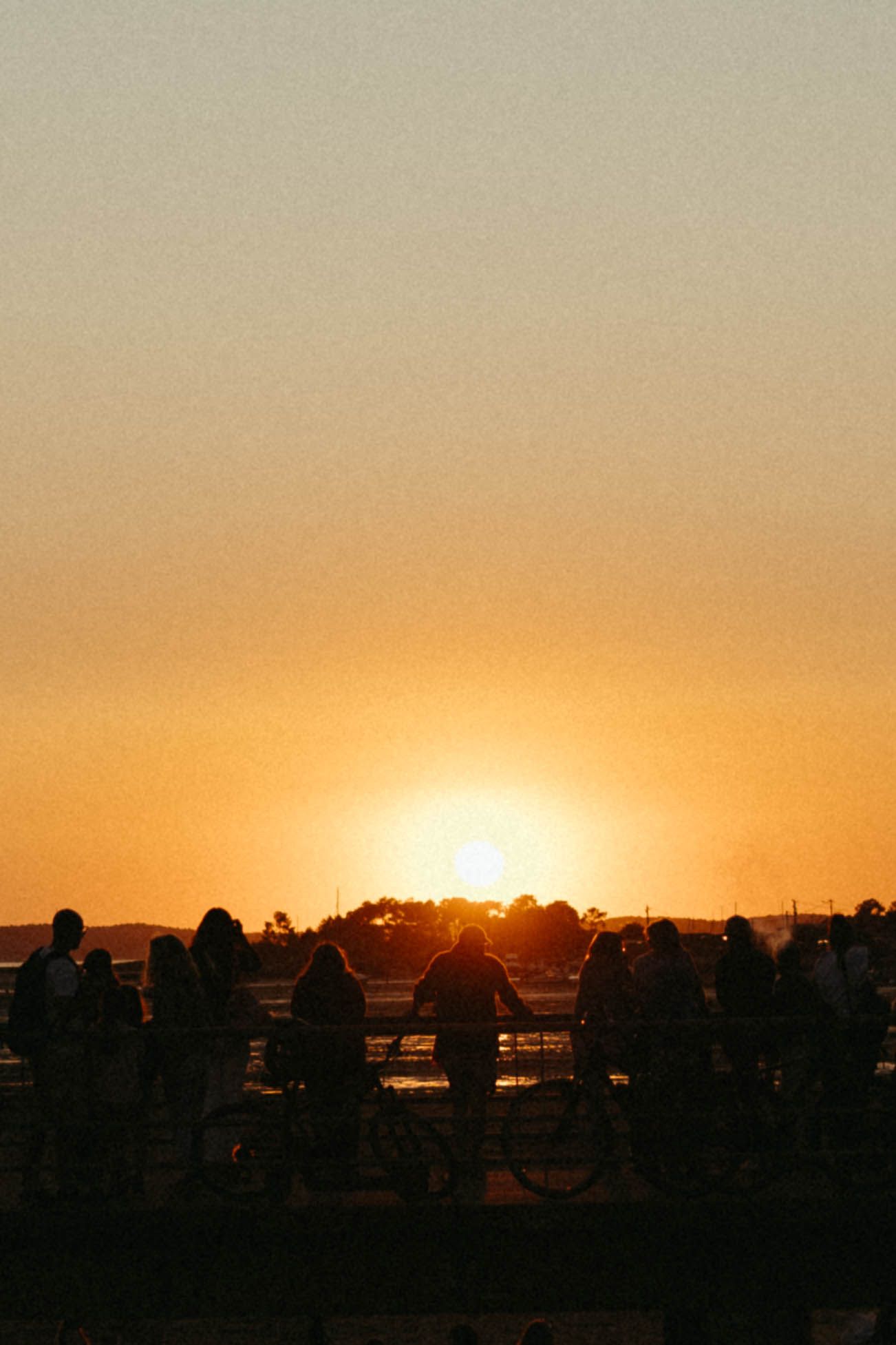 Silhouettes de personnes rassemblées au coucher du soleil, avec un soleil orange brillant à l'horizon contre un ciel clair.