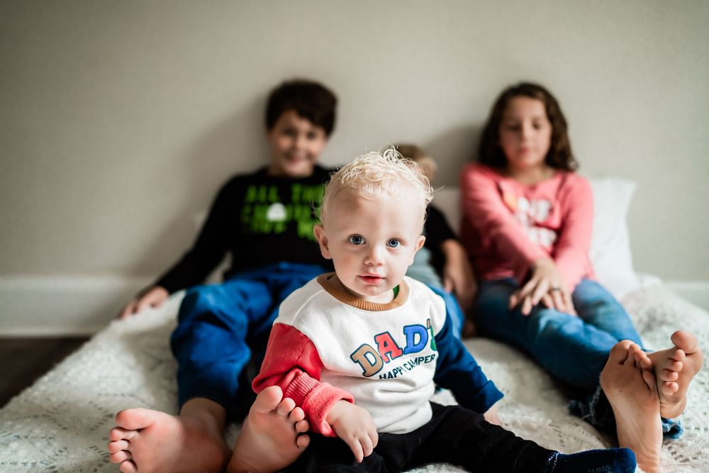 Three young children sitting together on a bed, with a blonde toddler in a white shirt in the foreground.