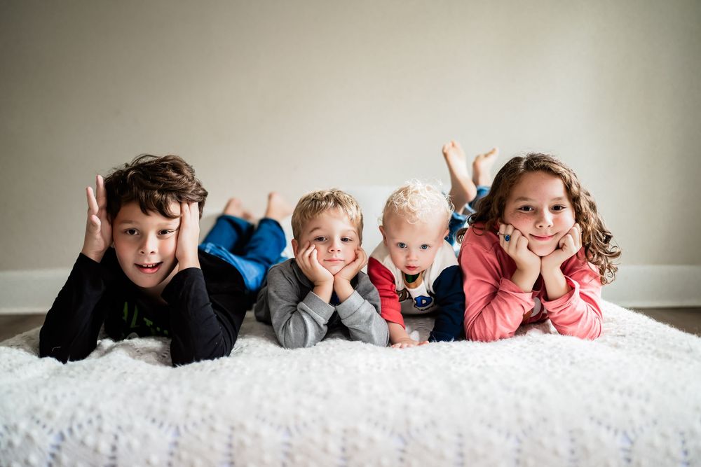 Four young children lying on a white fluffy rug, smiling and posing with hands on cheeks in a bright room.