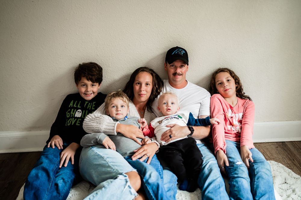 Family of six posing together on floor, parents with four young children smiling warmly against neutral wall.