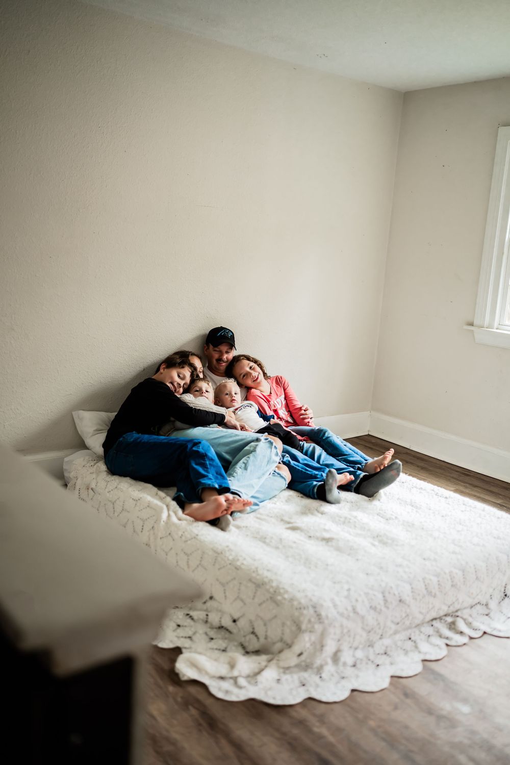 Family of four cuddling together on a white bed in a cozy bedroom with hardwood floors.