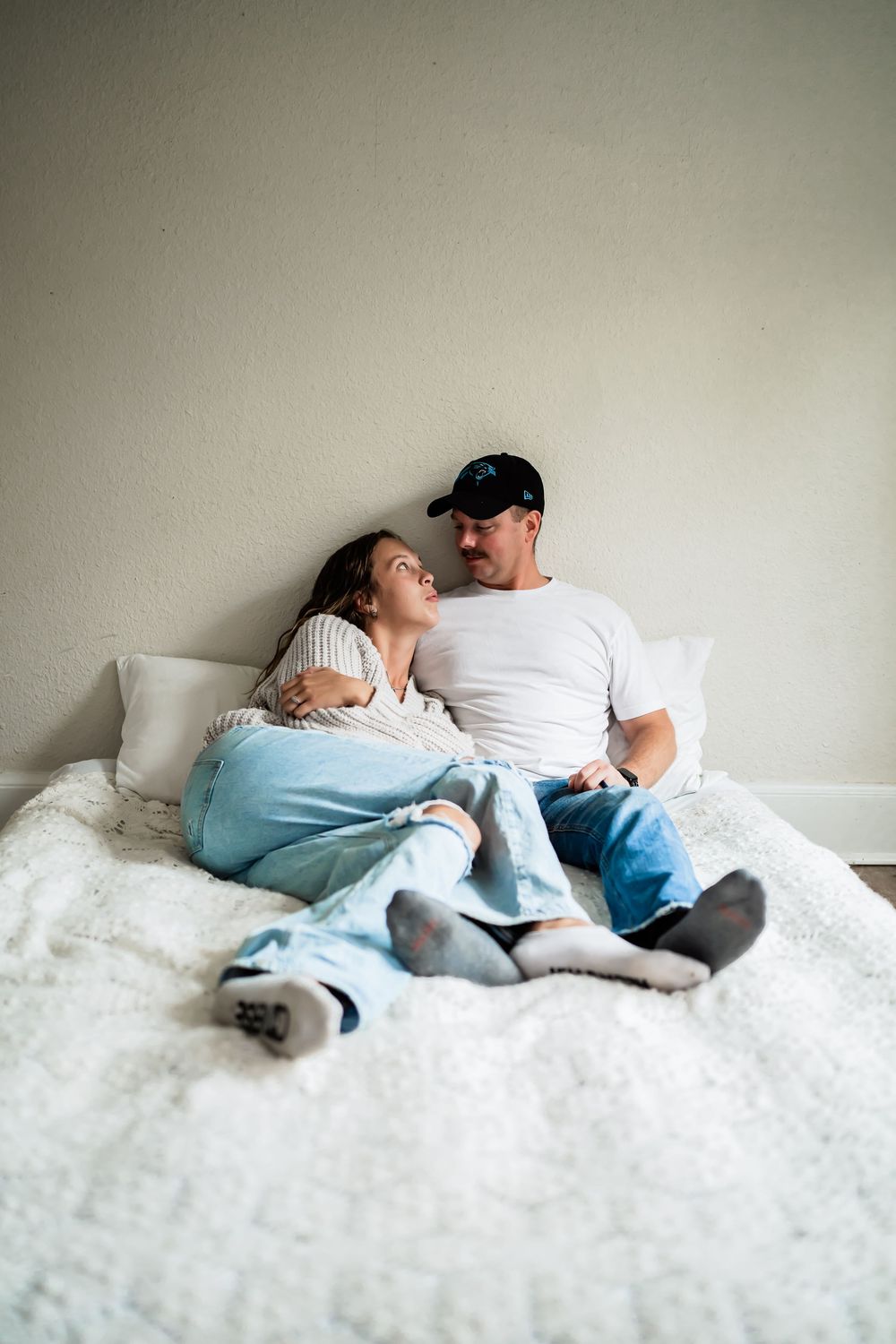 Couple relaxing together on a white fluffy bed, smiling at each other in a cozy bedroom setting.
