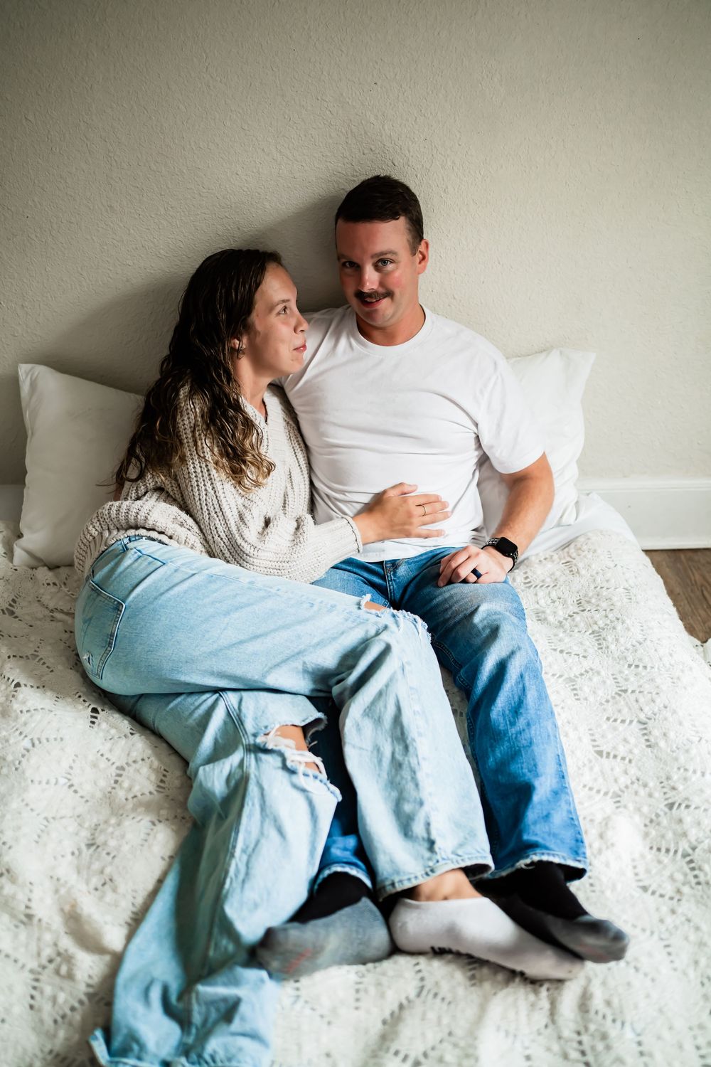 Couple sitting together on a white lace bedspread, wearing jeans and casual tops, sharing a warm moment.