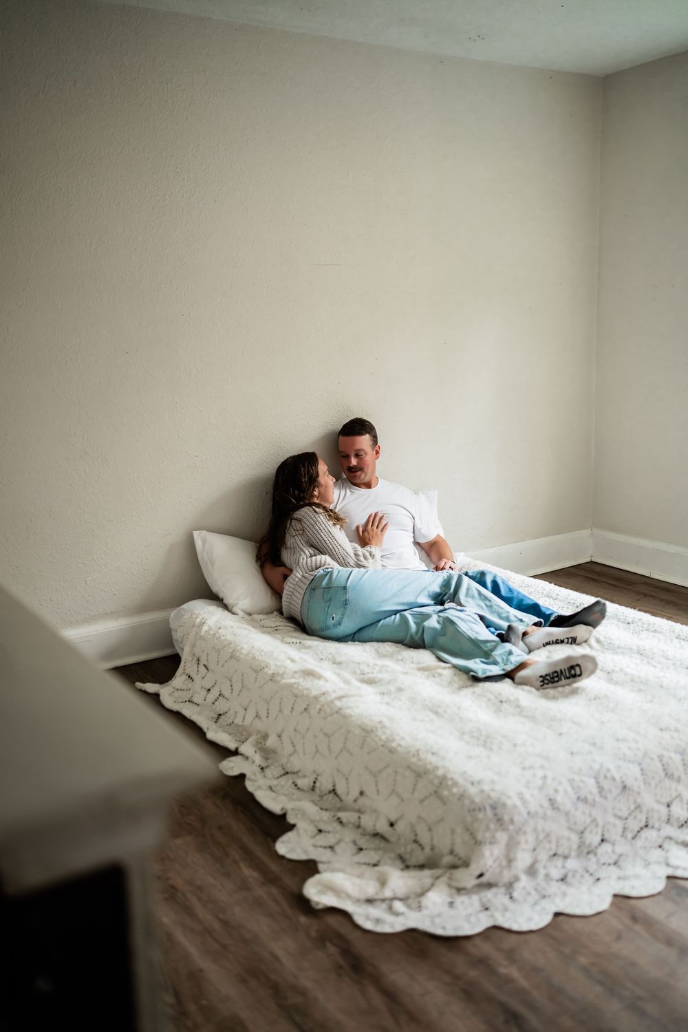 Couple cuddling on a white textured bed in a cozy bedroom with hardwood floors.