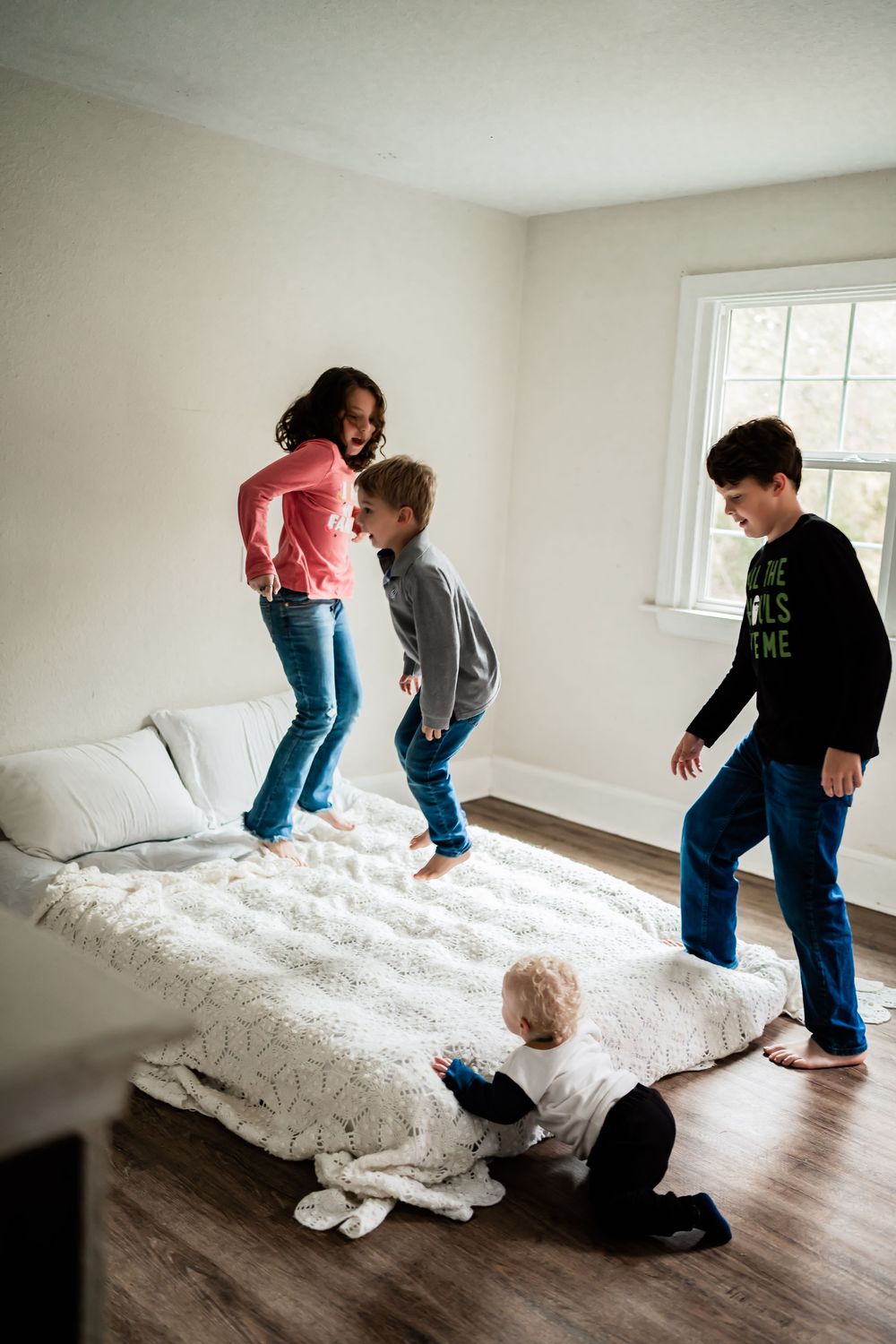 Three children jumping and playing on a white bed in a bright bedroom with hardwood floors.