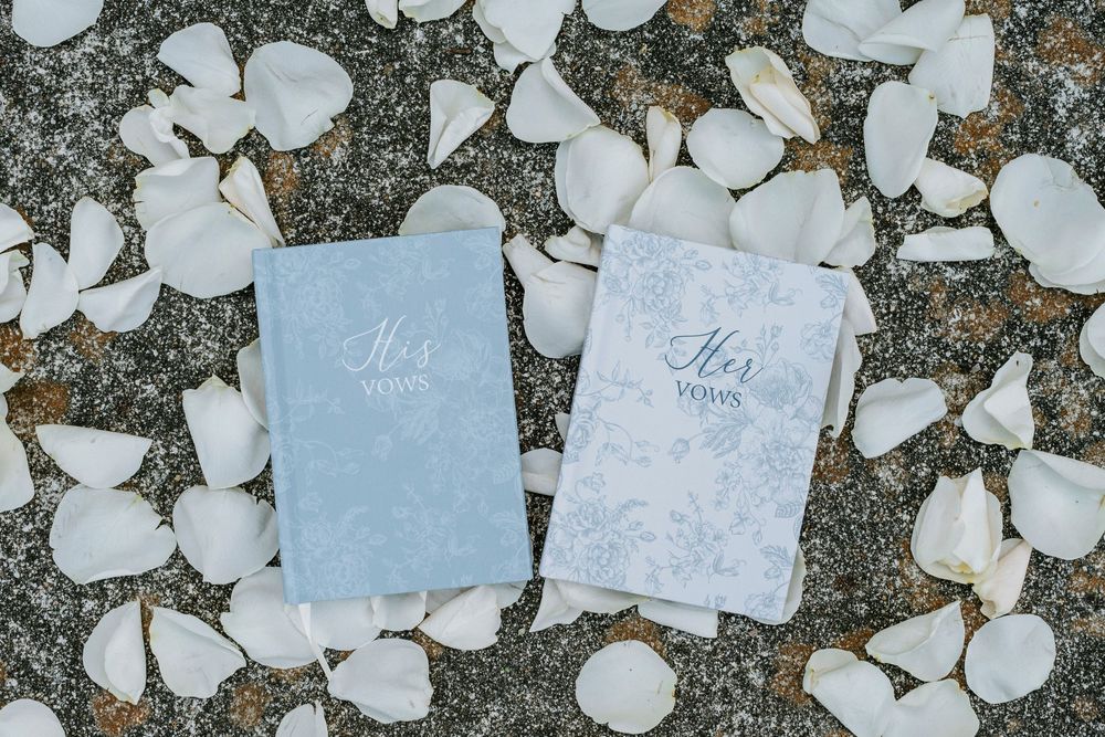 Two wedding vow books in light blue and white surrounded by scattered white rose petals on a stone surface.