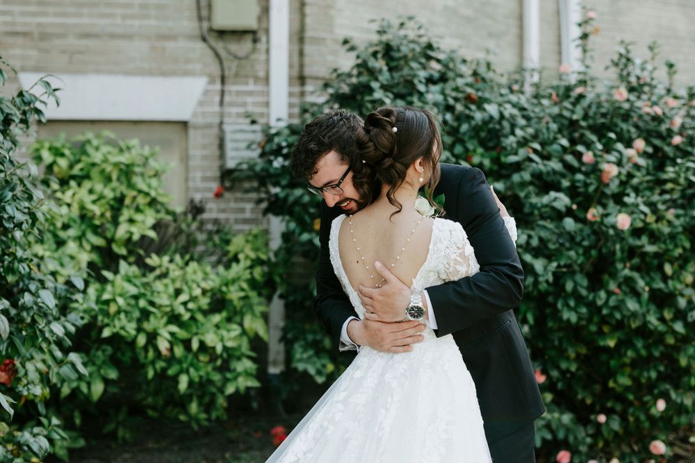 A couple in wedding attire embrace in a garden setting with lush greenery and brick walls in the background.