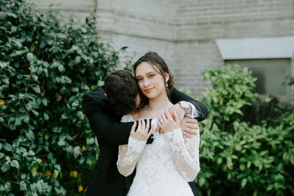 A couple embraces during their wedding ceremony in front of lush green foliage.
