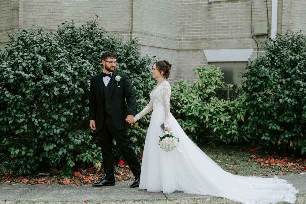 A wedding couple walks through a garden pathway with greenery and white walls creating an elegant outdoor setting.