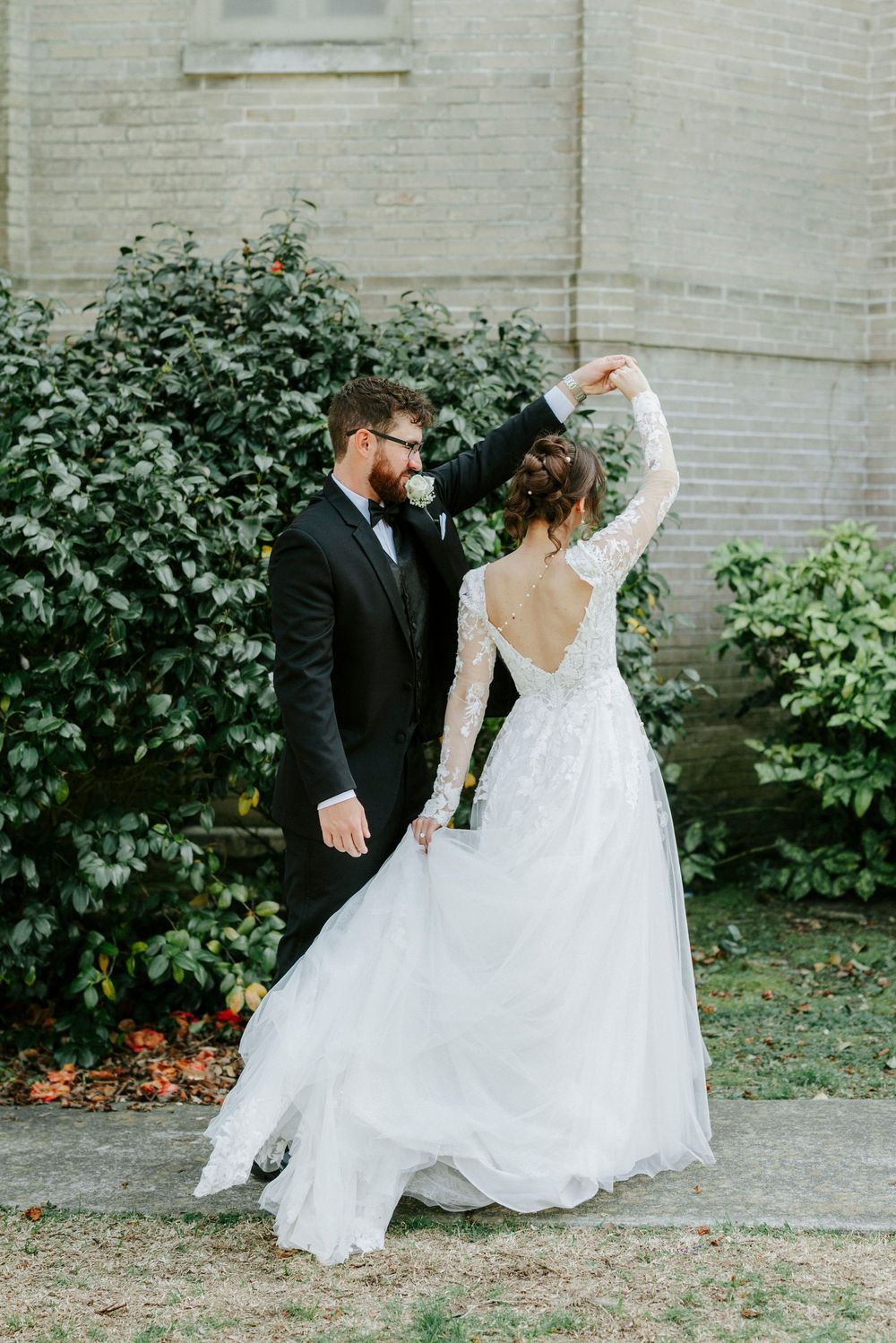 A couple in wedding attire dances and twirls together in front of a brick wall and garden setting.