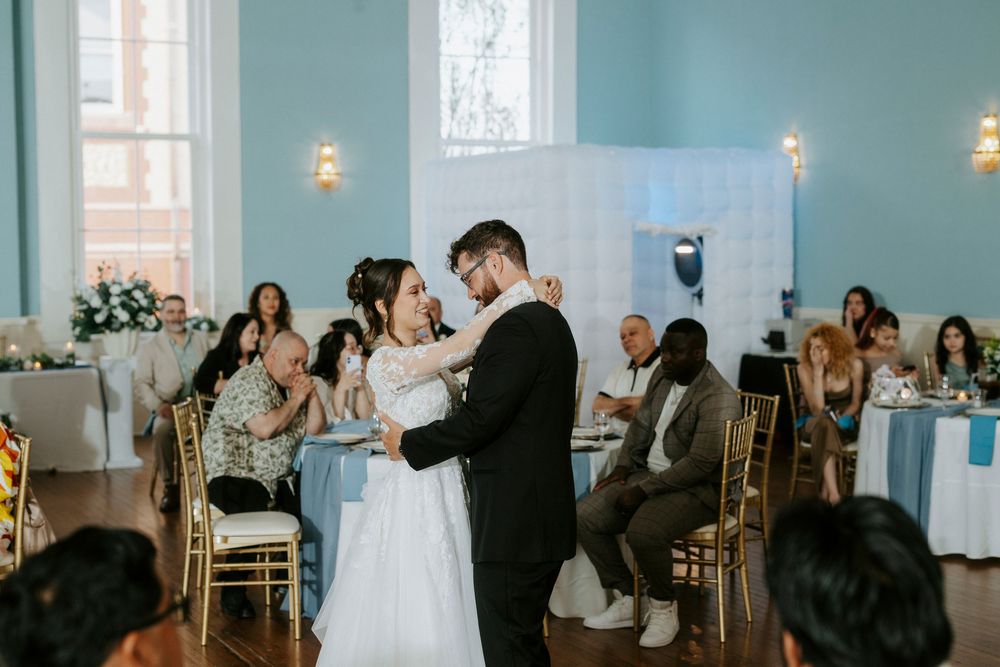 A couple shares their first dance surrounded by guests at an elegant reception with gold chairs and floral decor.