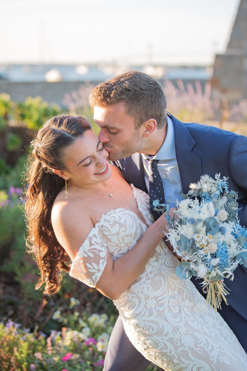 Romantic wedding couple shares intimate moments at sunset in a garden with blue floral bouquet.