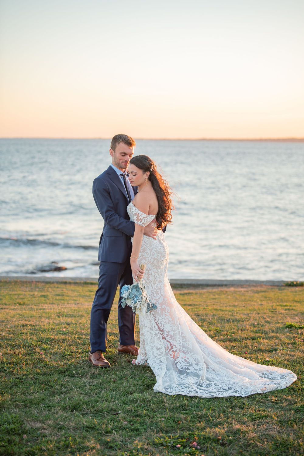 A romantic seaside wedding portrait with a couple embracing on grassy cliff overlooking ocean at sunset.