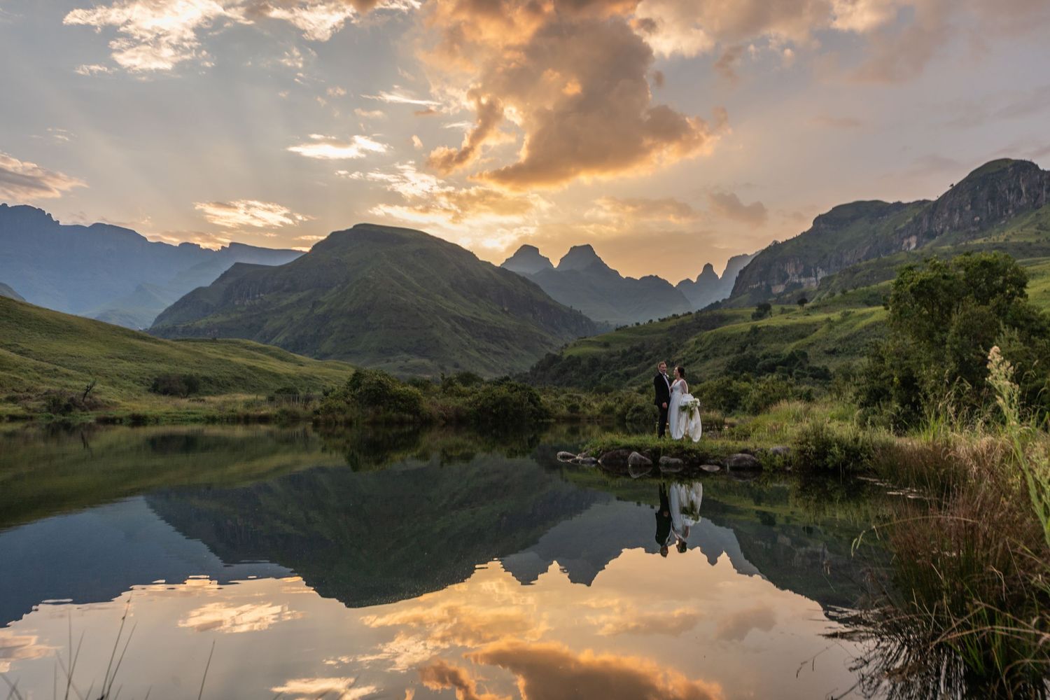 Couple embracing by a reflective mountain lake at sunset with dramatic clouds and lush green valleys in the background.