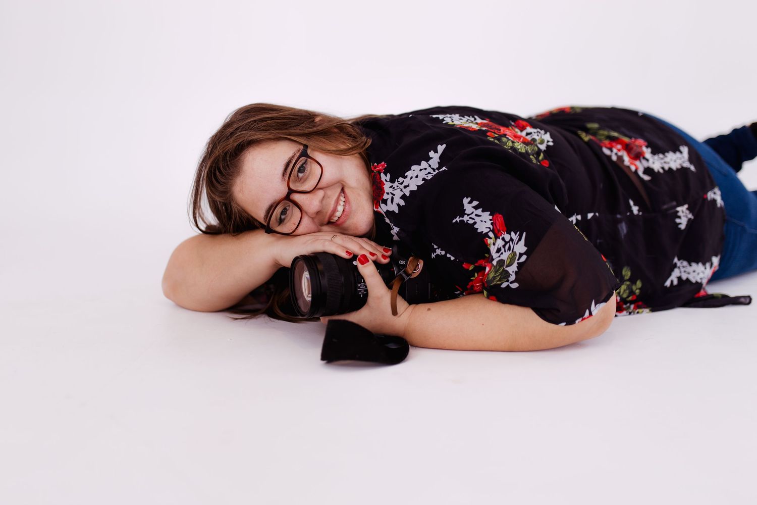 Thalie photography female photographer lying on white background holding a professional DSLR camera, wearing floral shirt and glasses.