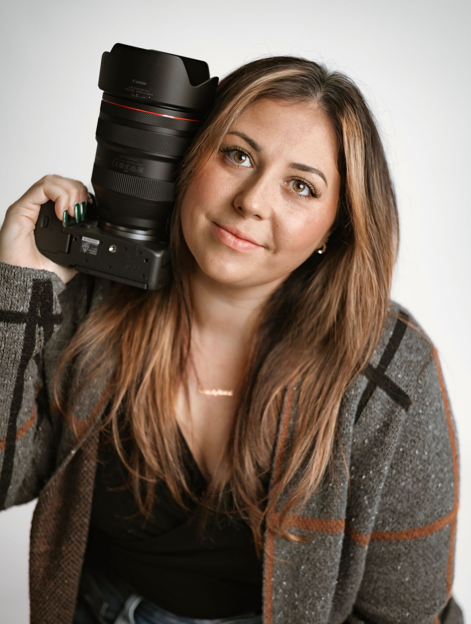 Female photographer holding Canon DSLR camera on shoulder against gray background.