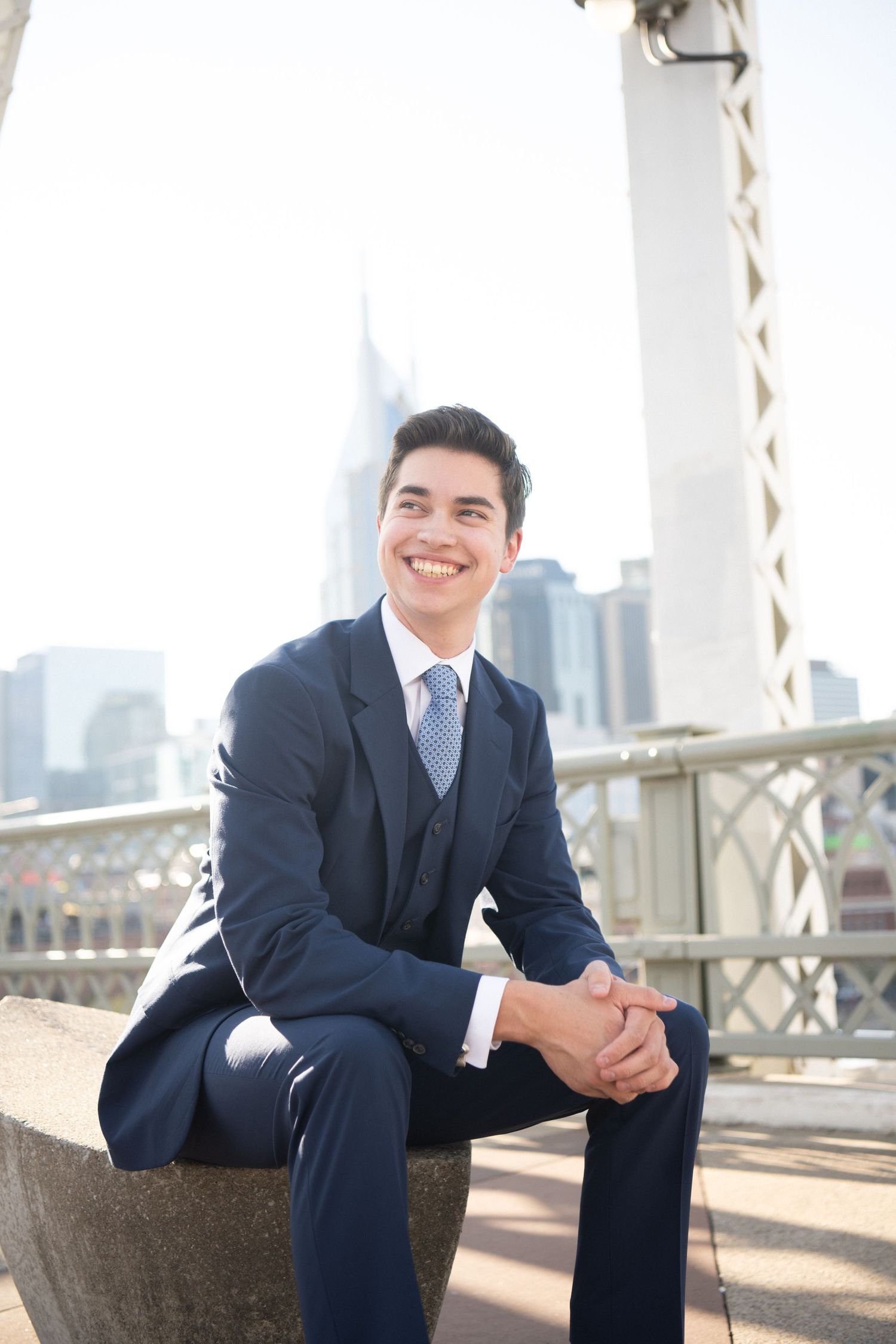 A businessman in a dark suit sits thoughtfully on a concrete ledge with a cityscape backdrop on a sunny day.