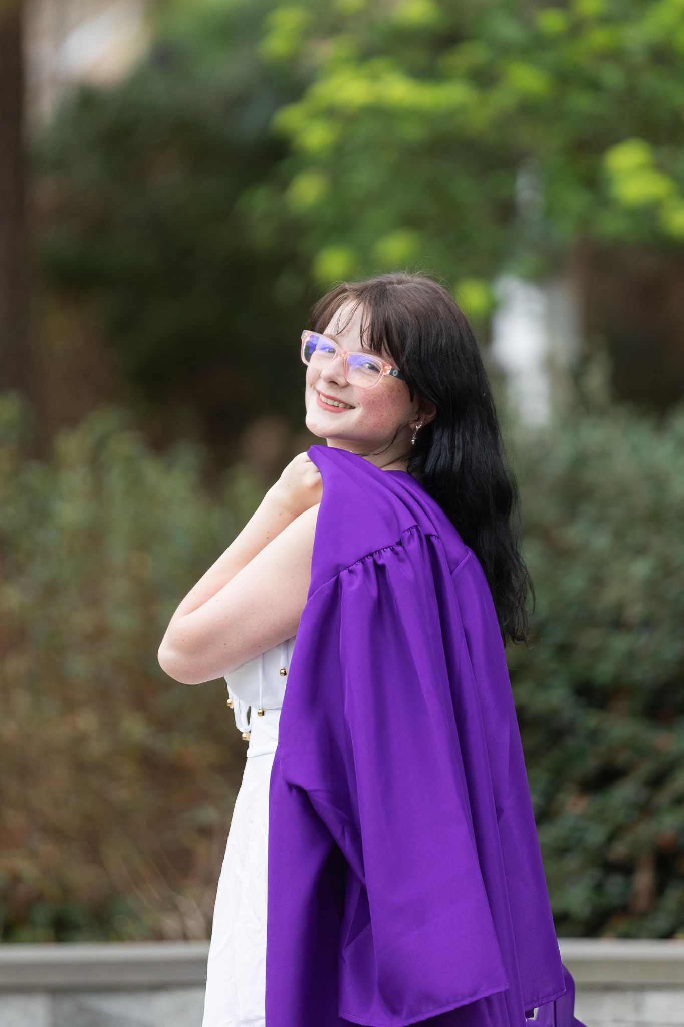 Young woman in white dress holding purple graduation gown, smiling over shoulder outdoors at WRAL Azalea Garden