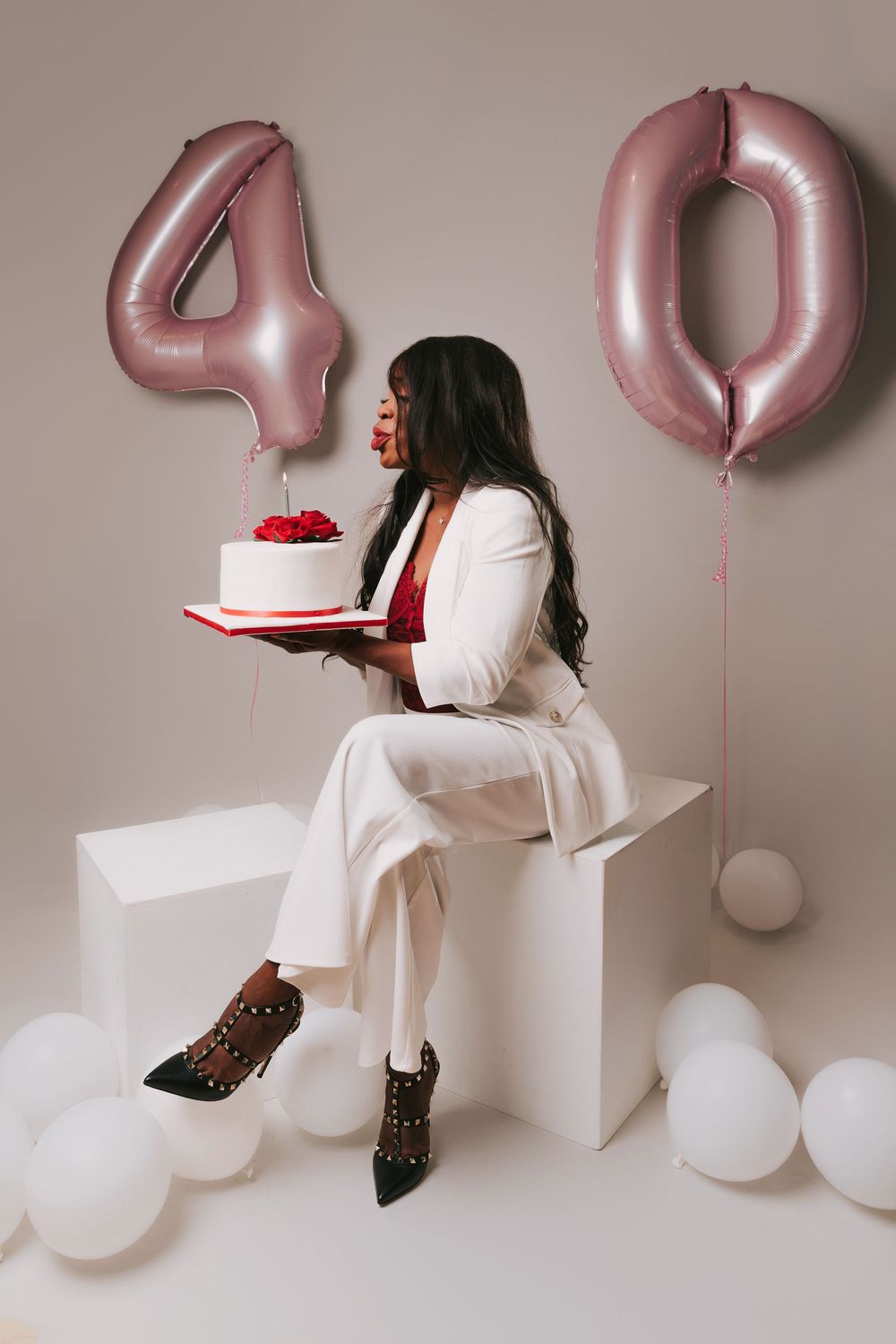 Elegant birthday celebration setup with rose gold number 40 balloons, white decor, and a person holding a red-topped cake.