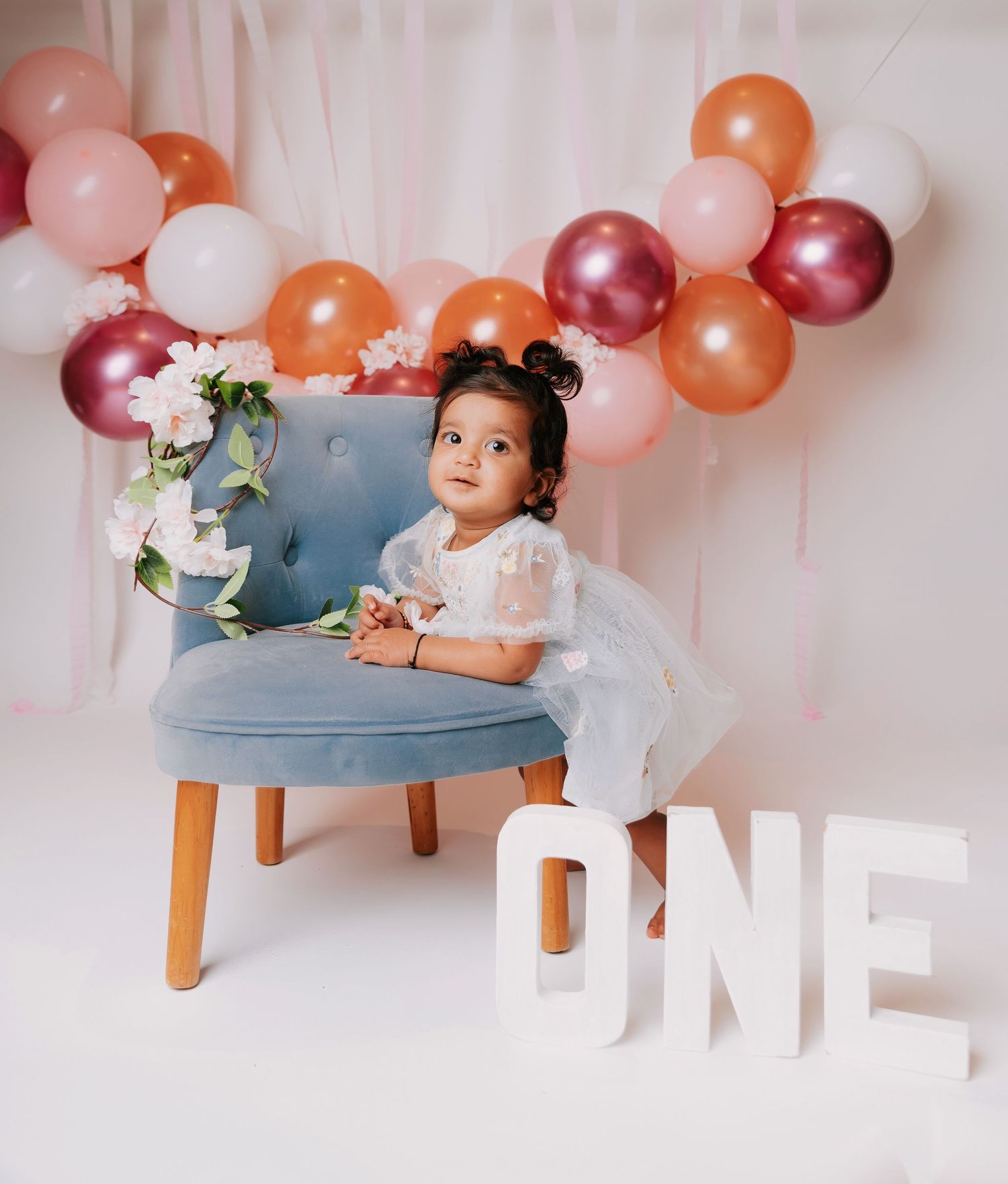 A baby in a white dress sits on a blue chair under pink and orange balloons for a first birthday photoshoot.