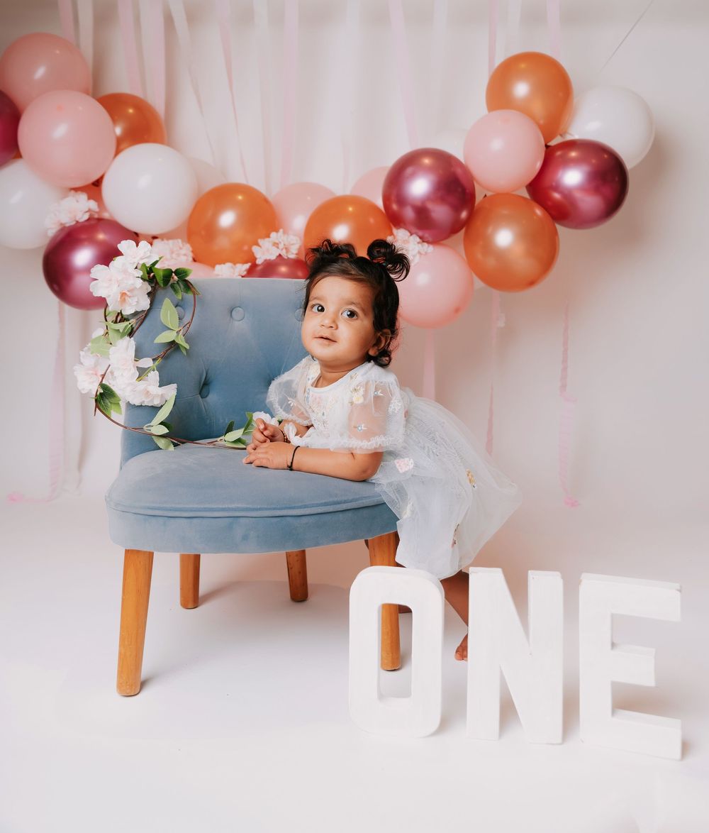 A baby in a white dress sits on a blue chair under pink and orange balloons for a first birthday photoshoot.
