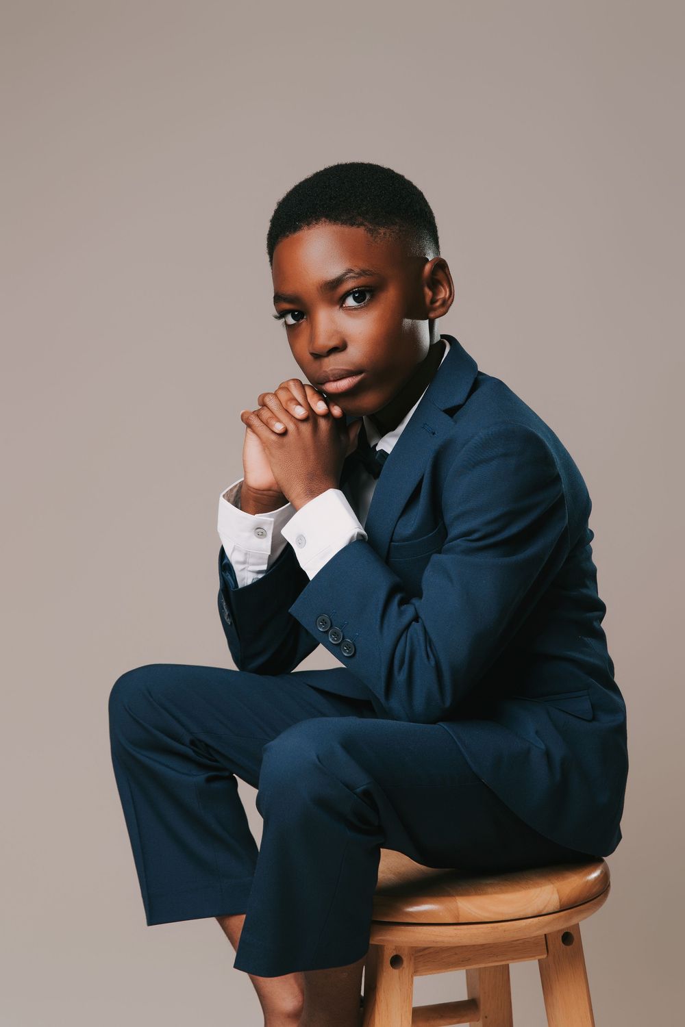 Professional studio portrait of someone in a navy blue suit sitting thoughtfully on a wooden stool against neutral backdrop.