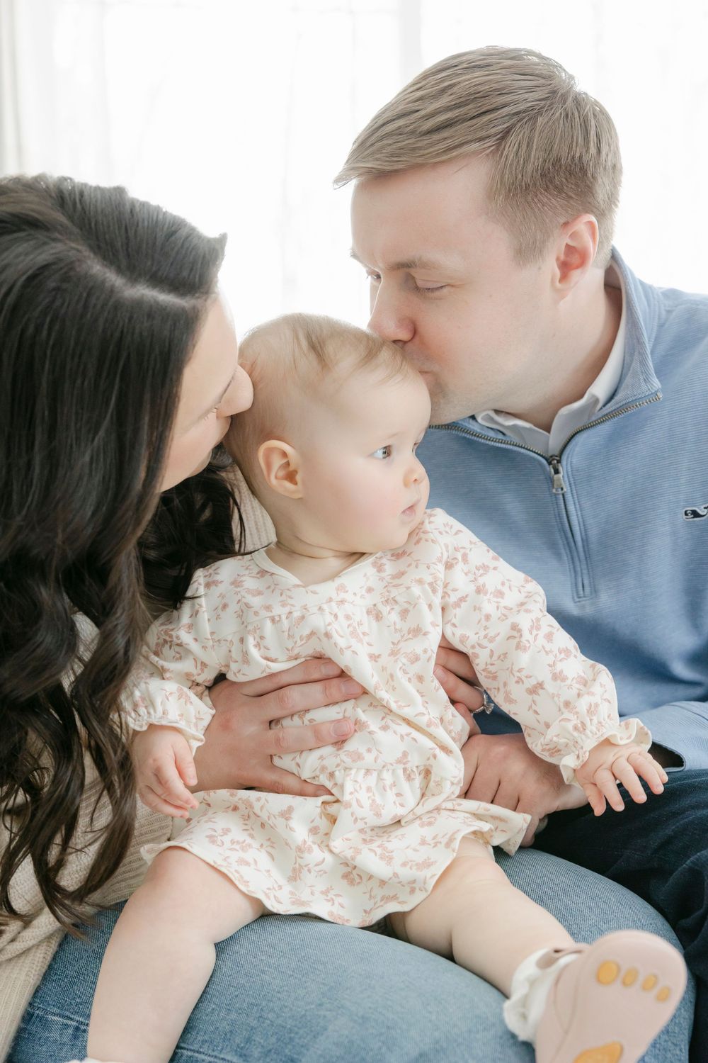 A tender family moment shows parents sharing affection with their baby wearing a floral dress.