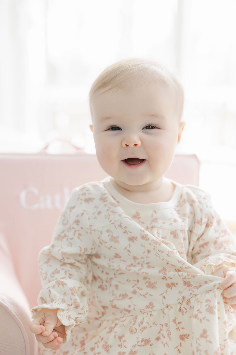A baby in a floral onesie smiles and laughs while laying on white bedding in a bright room.