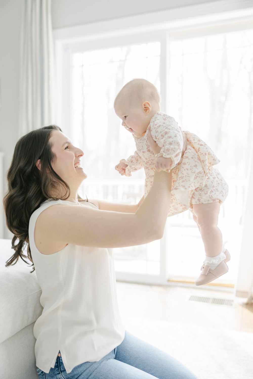 A parent lifts and plays with a baby in a bright sunlit room against white curtains.