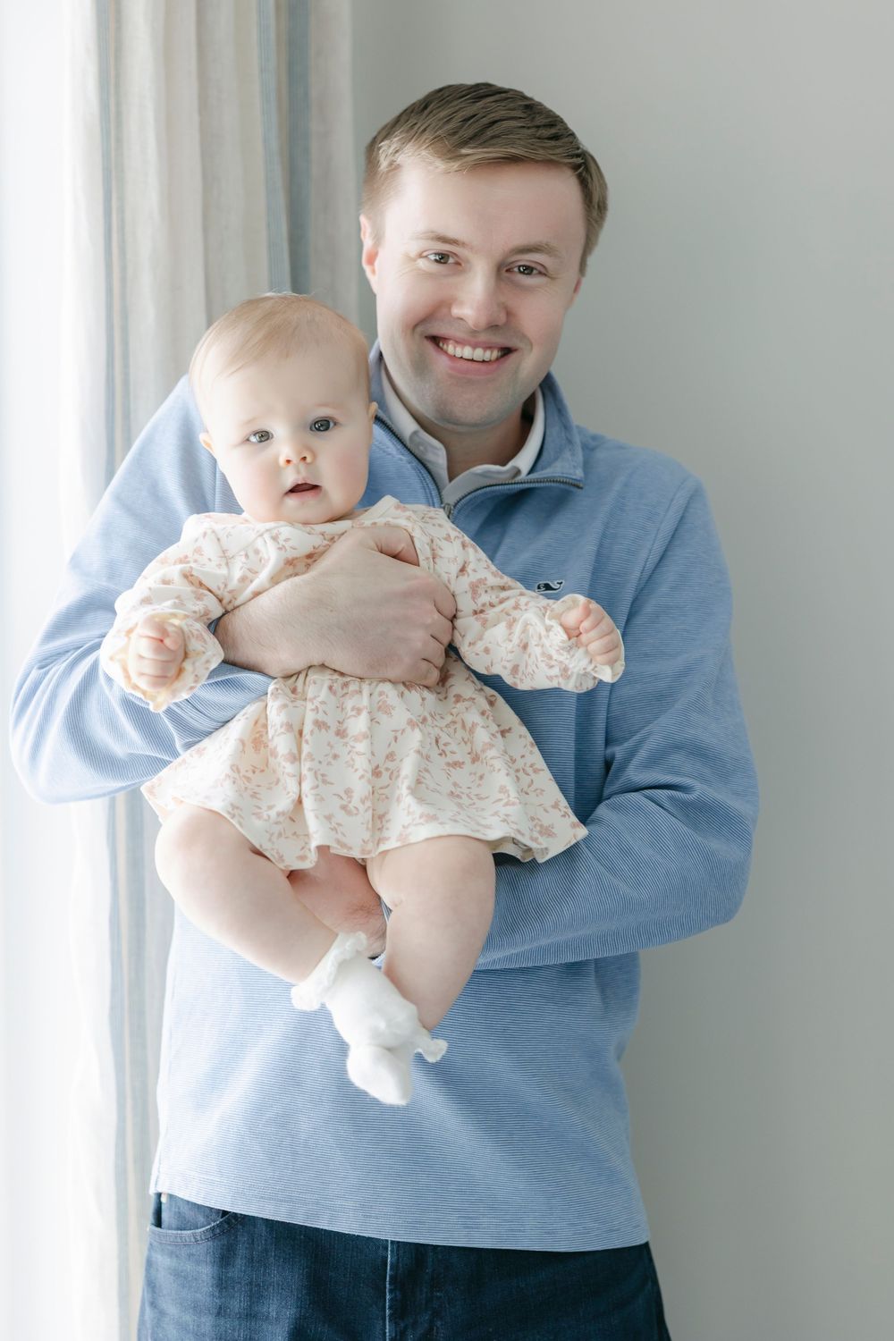 A person in a blue sweater holds a baby wearing a light-colored dress against a white background.