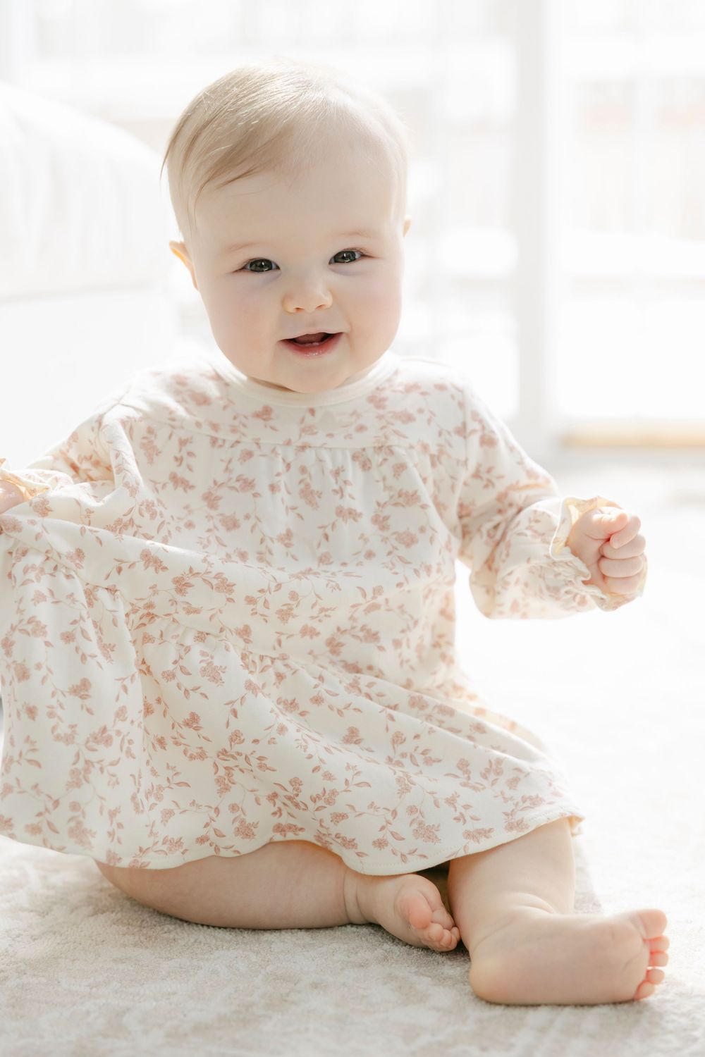 A smiling baby in a floral dress crawls and plays on a white floor near a bright window.