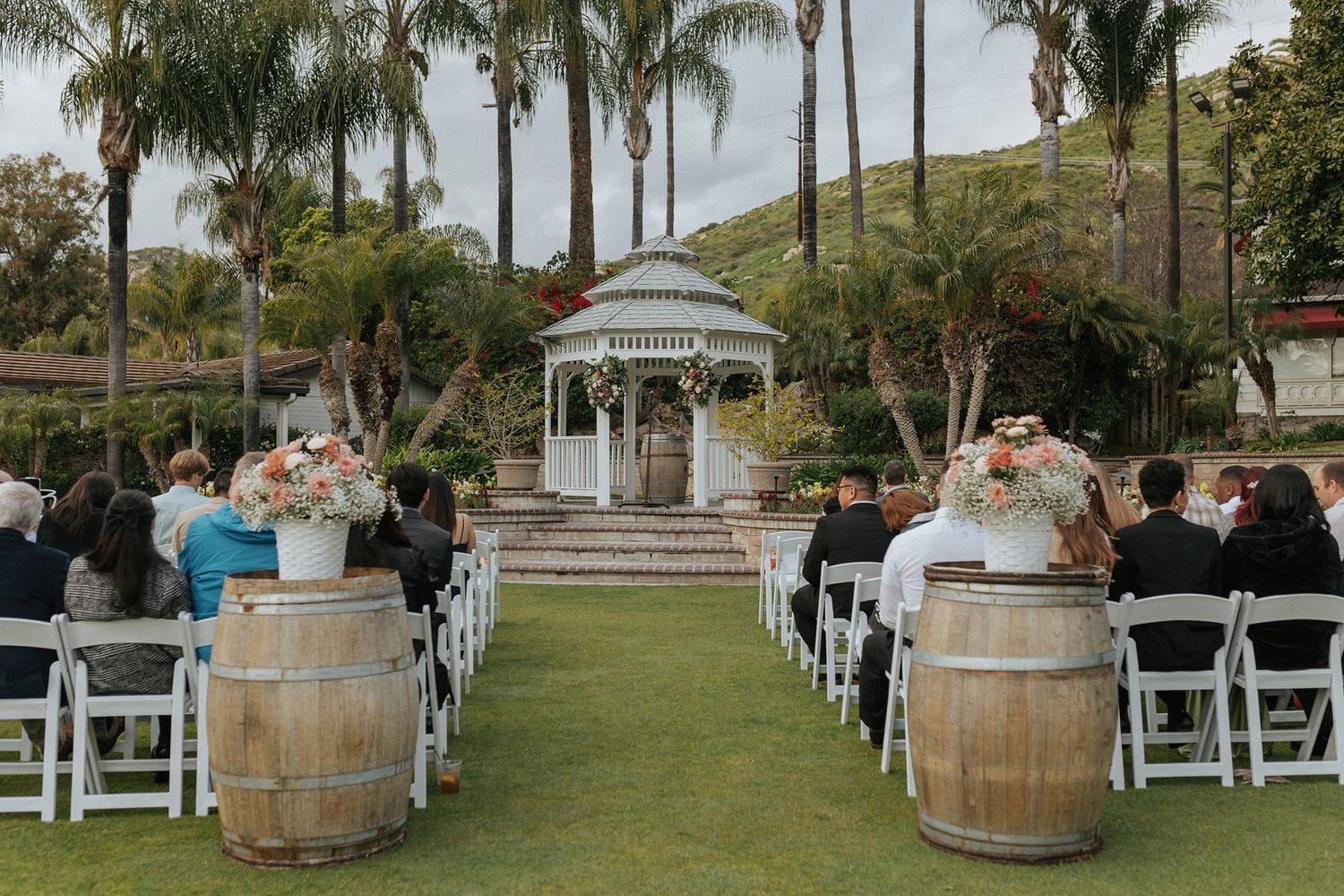 Panoramic view of an outdoor wedding ceremony setup with a gazebo, white chairs, and wine barrels along a palm tree-lined aisle.