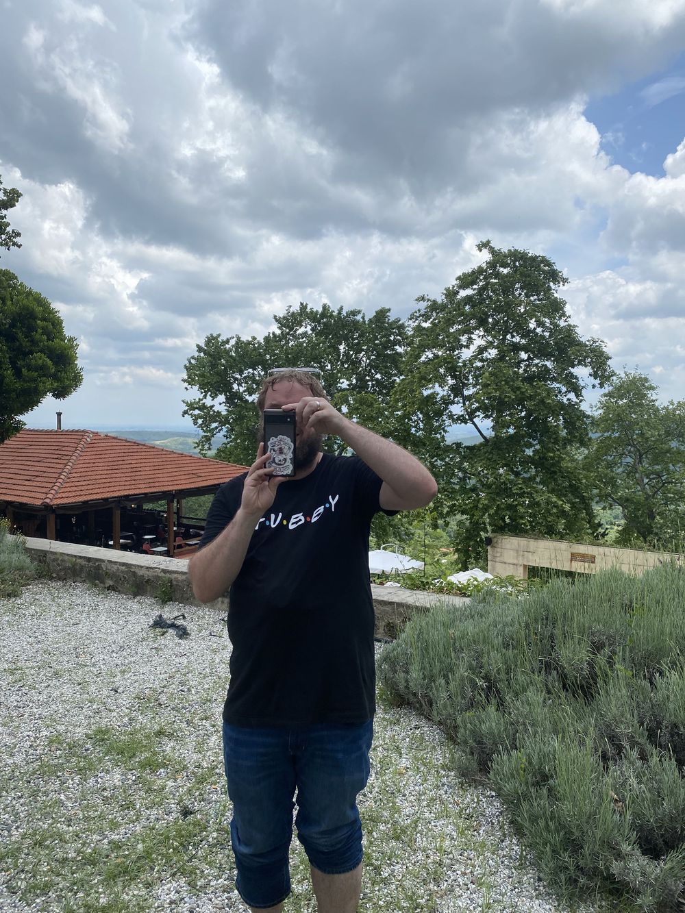 Someone in a black t-shirt and jeans takes a photo outdoors on a gravel path with trees and cloudy sky in background.