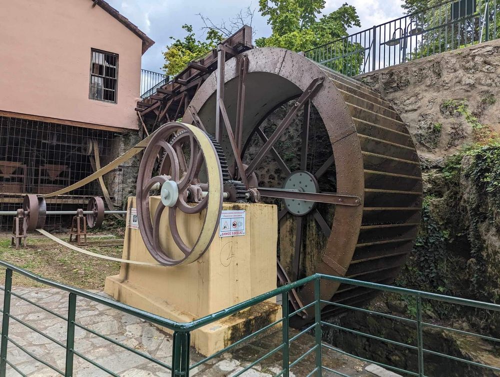 Historic water wheel and mill mechanism with stone walls and green railings along a walkway in an outdoor setting.