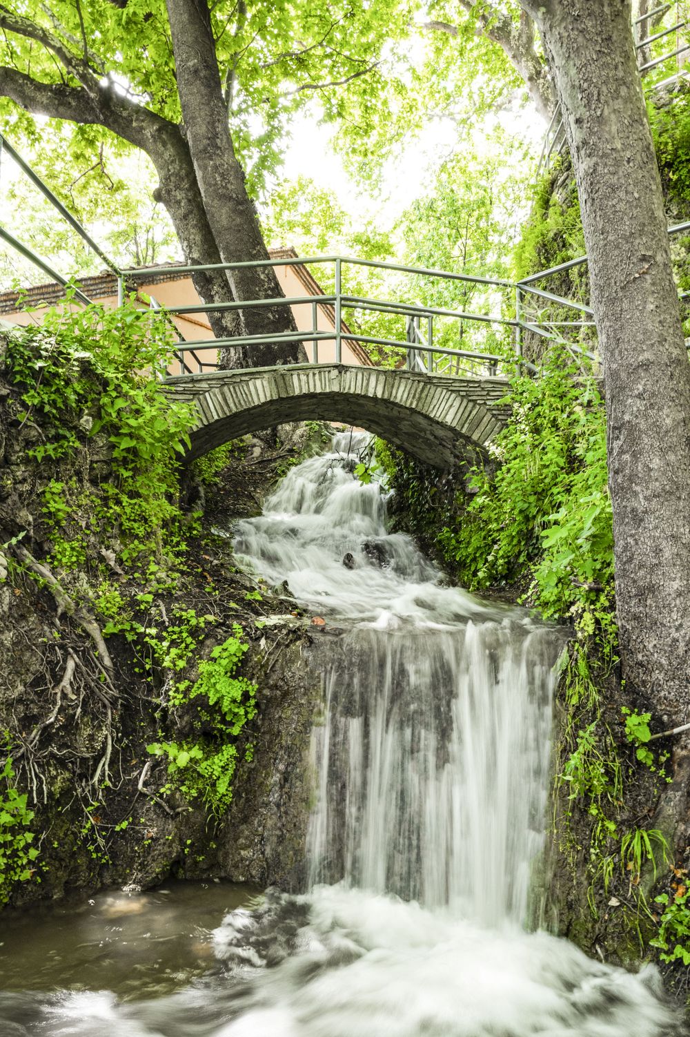 Stone bridge arches over a cascading waterfall surrounded by lush green foliage in a peaceful forest setting.